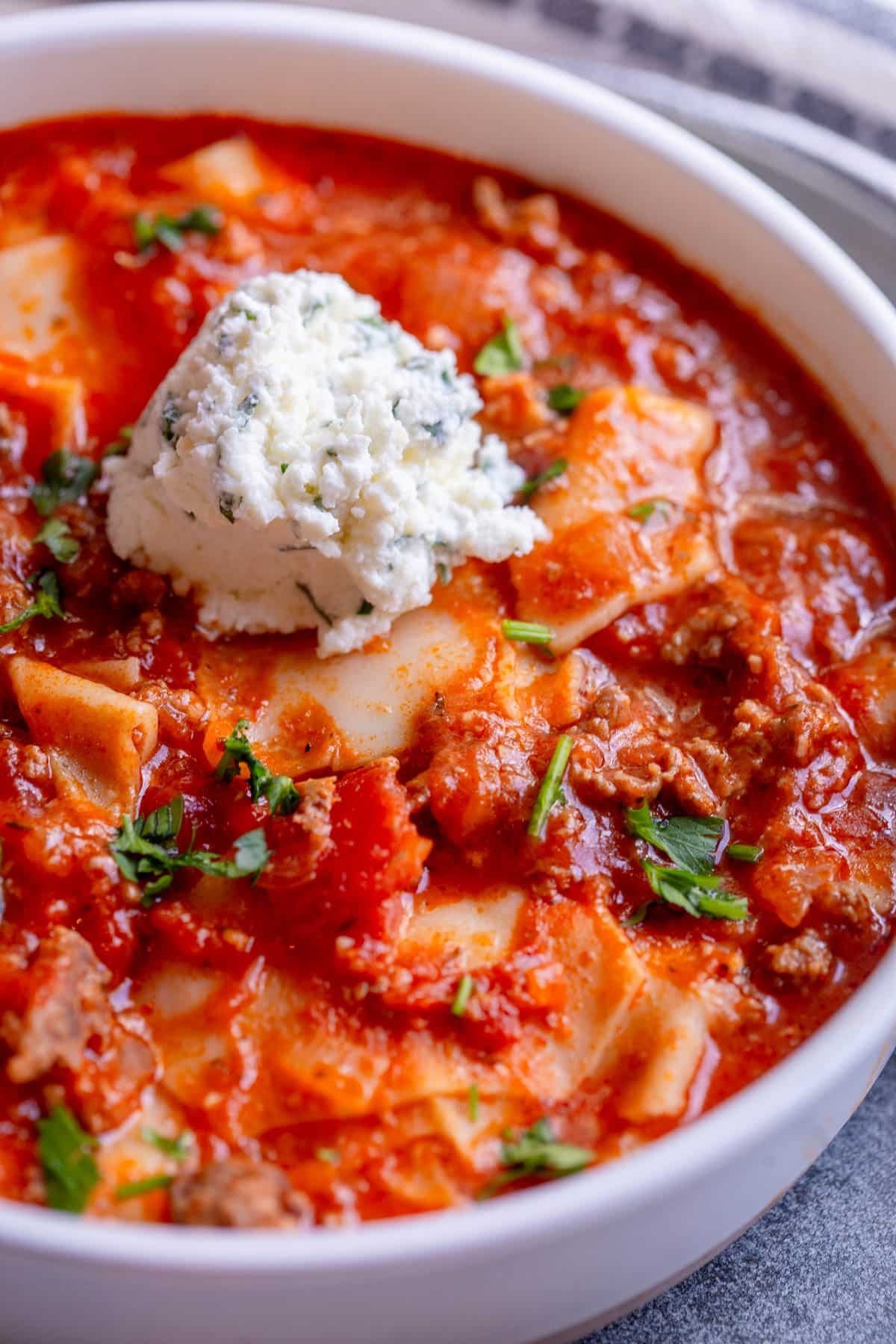 Slow Cooker Lasagna Soup in a shallow cream colored bowl with a dollop of ricotta on top and sprinkle of fresh parsley. 