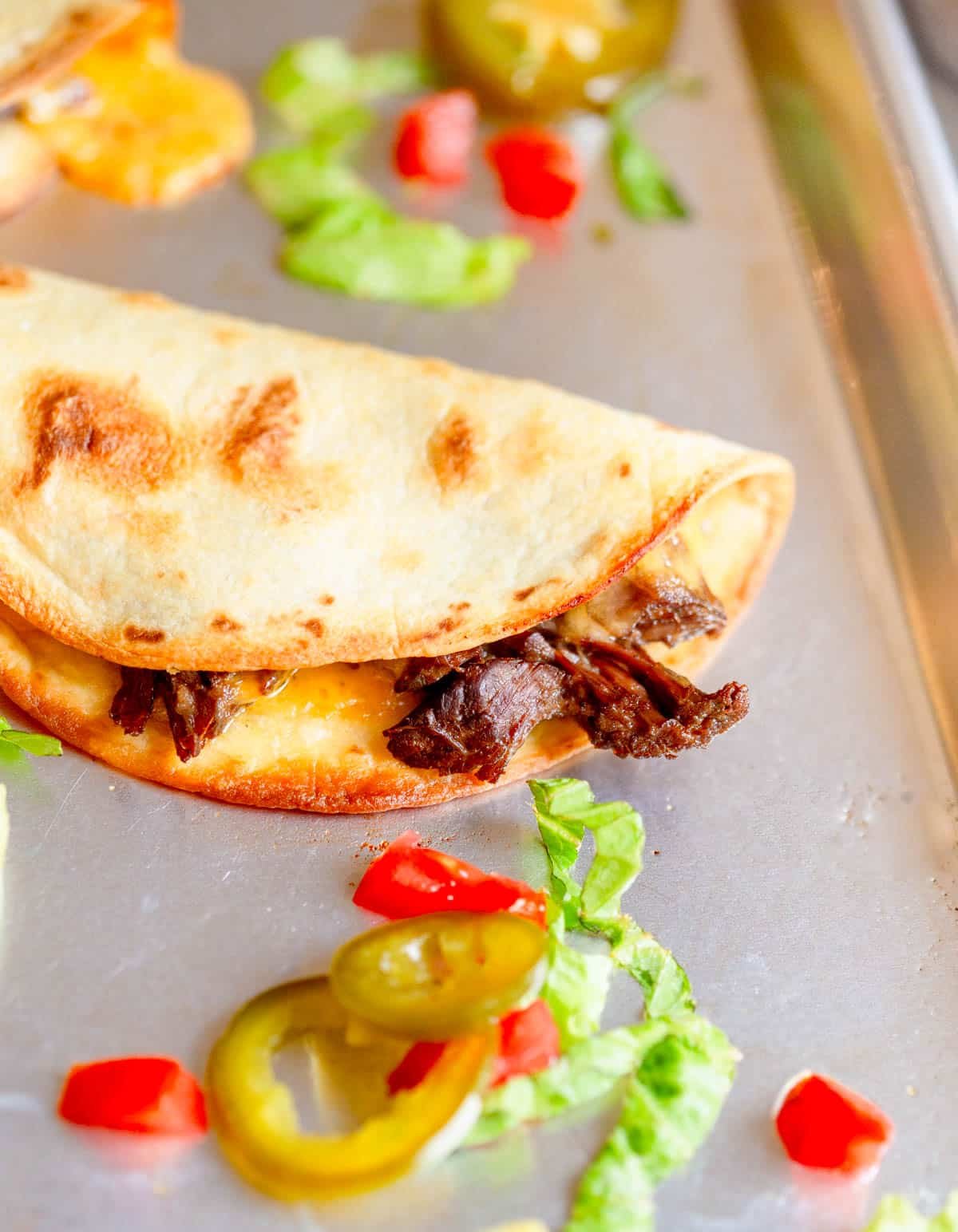 Slow Cooker Shredded Beef Tacos on a stainless steel baking sheet with shredded lettuce, pickled jalapeños, and diced tomatoes around it.