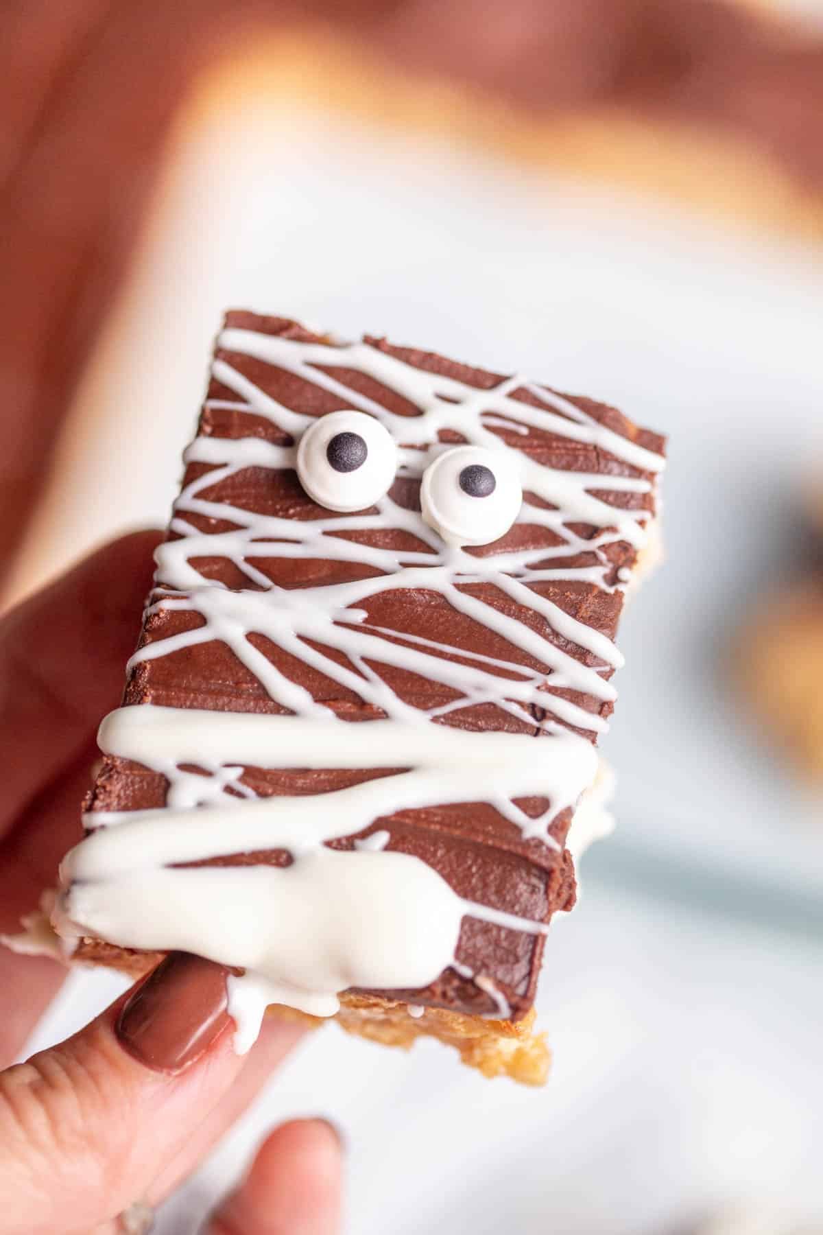 A hand with brown finger nails holding a Halloween Scotcharoo drizzled with white chocolate and decorated with edible eyes. 