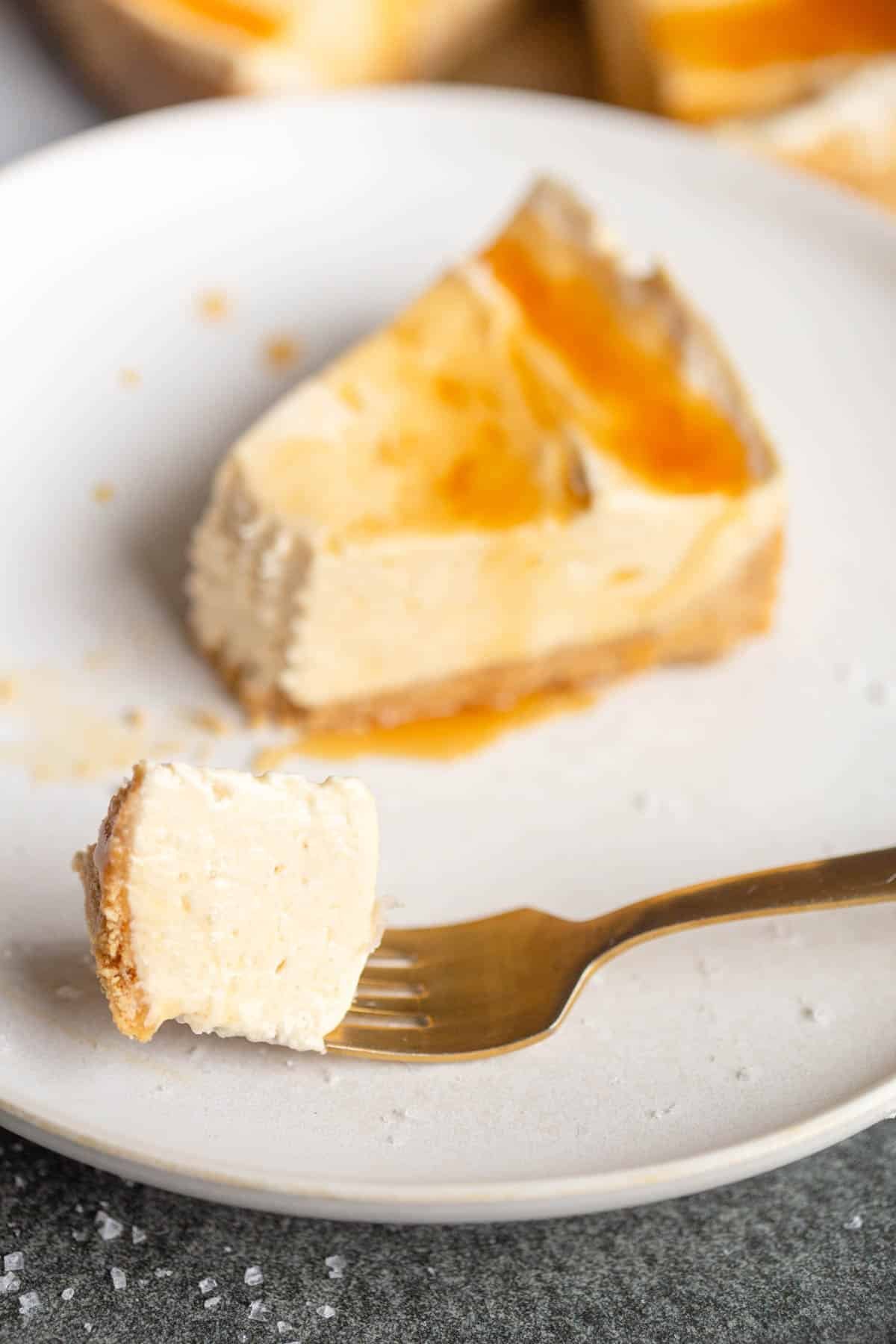 A slice of no bake salted caramel cheesecake on a white plate with a gold fork topped with a drizzle of caramel and sea salt. 