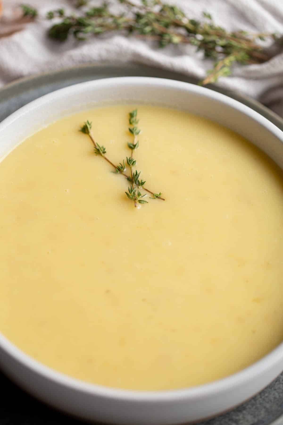 Slow Cooker Potato Leek Soup in a white bowl topped with two sprigs of fresh thyme, on a dark gray plate with a loaf of bread and more fresh thyme in the background. 