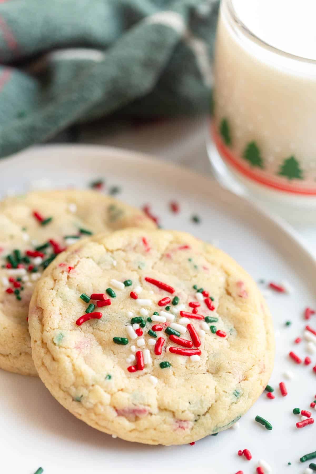 Christmas Sprinkle Sugar Cookies on a white plate with a mug of milk and a festive towel in the background. 
