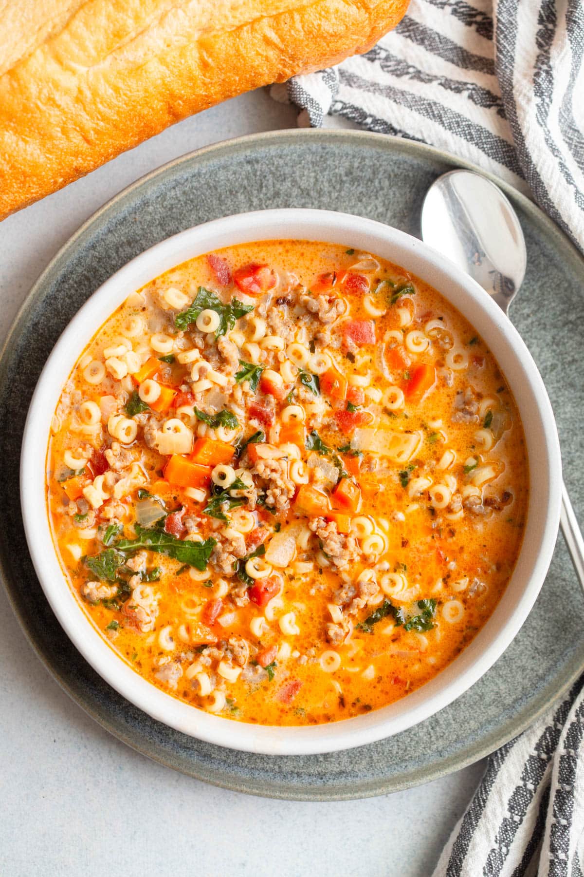 Creamy Italian Sausage Pasta Soup in a shallow cream colored bowl on a dark gray plate with a loaf of bread, a striped napkin next and spoon to it.