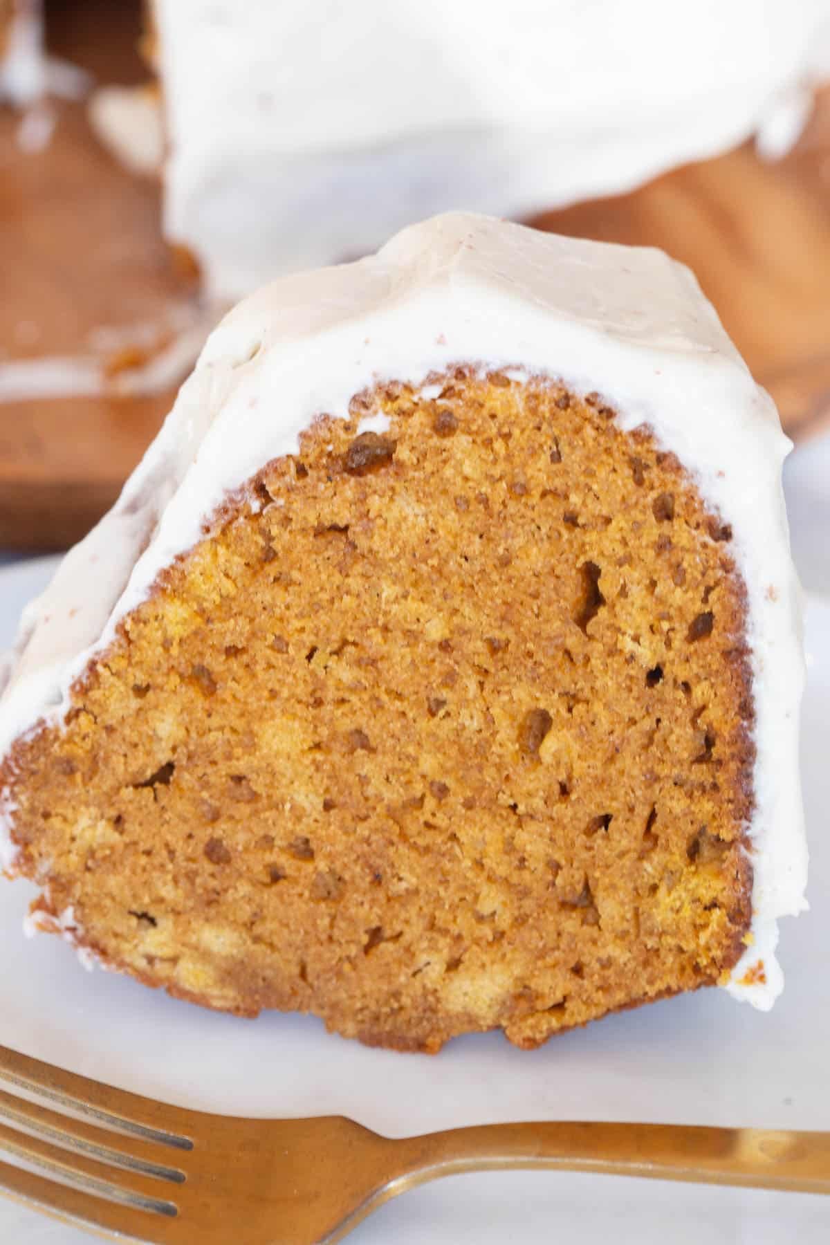 A slice of Pumpkin Bundt Cake with Brown Butter Frosting on a white plate with a gold fork in front of it.