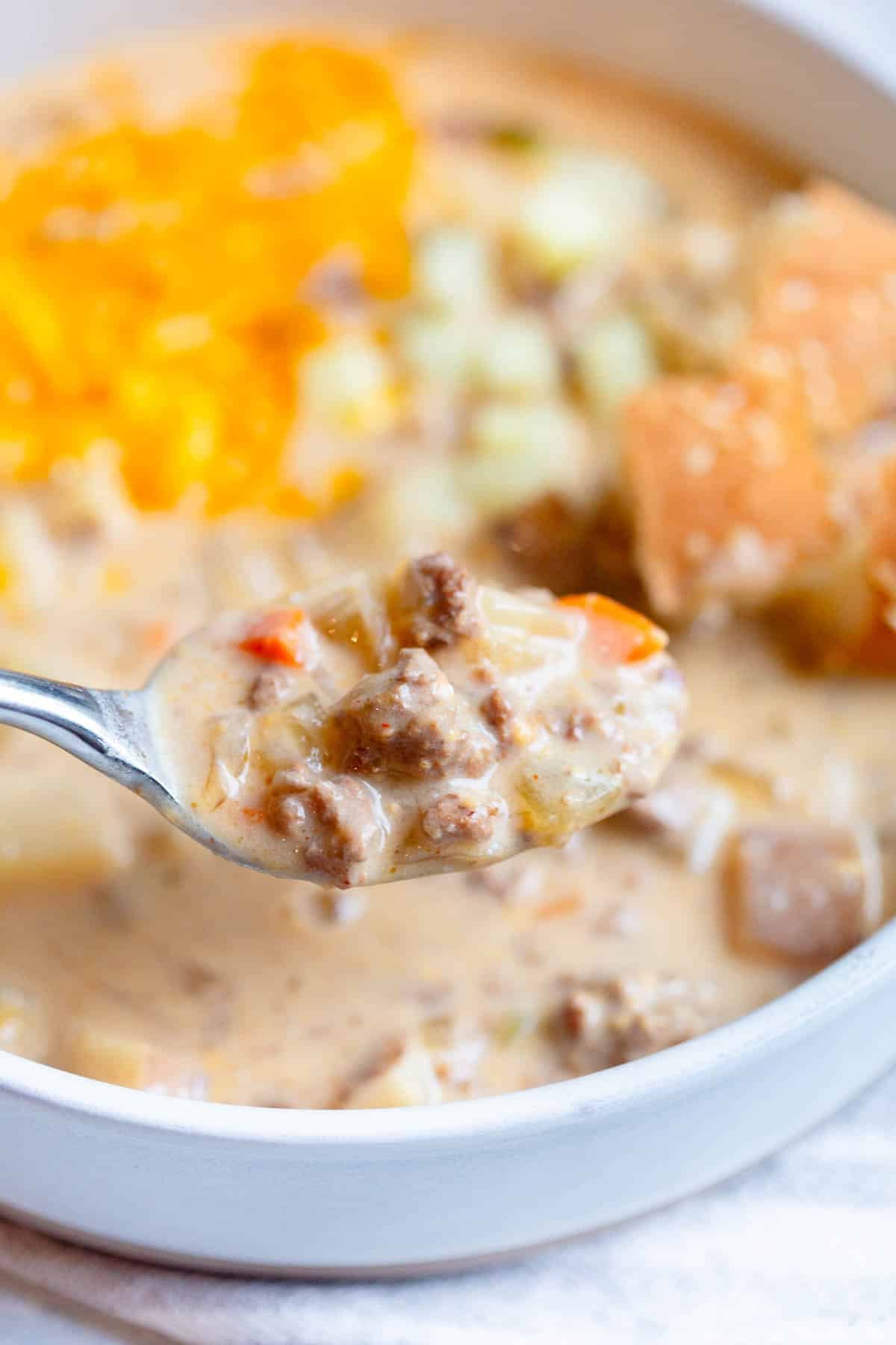 A silver spoon with a scoop of Slow Cooker Cheeseburger Soup, with the bowl of soup in the background.