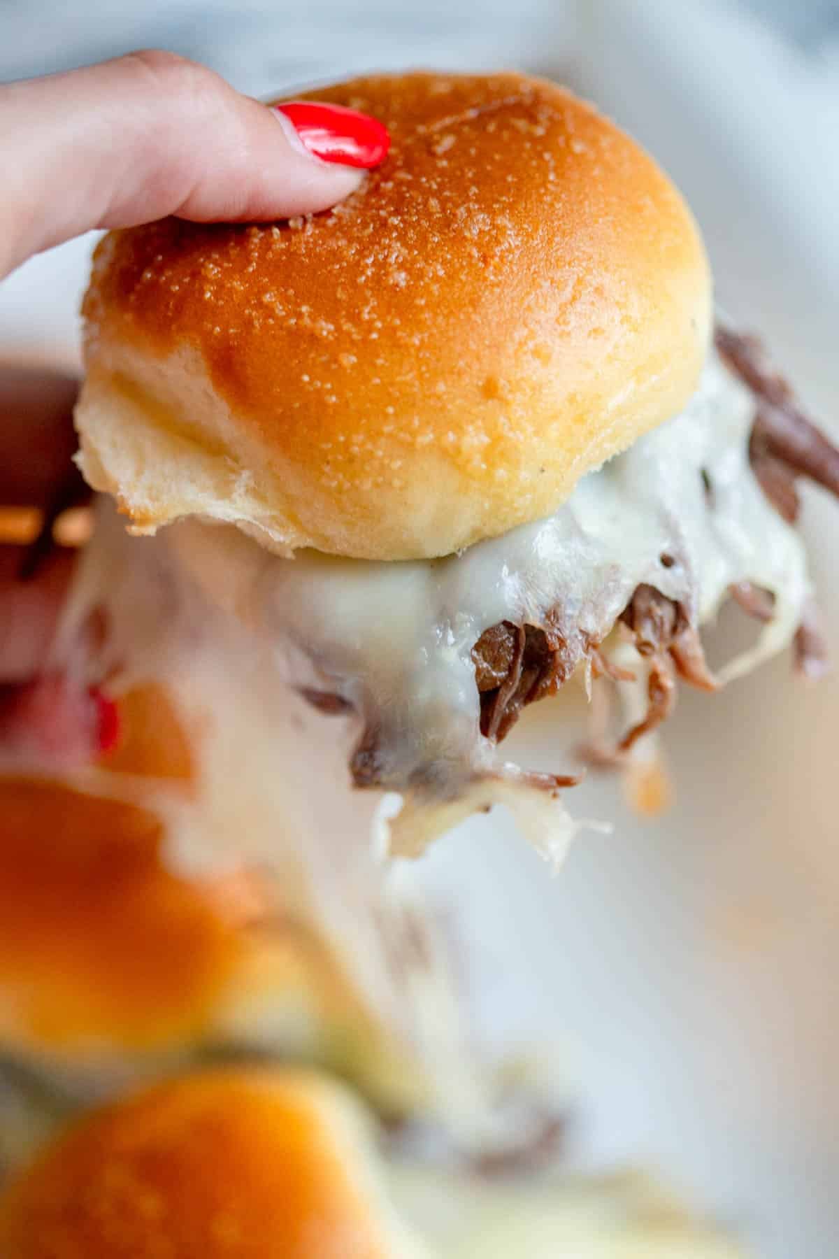 A hand with red fingernail polish holding a Slow Cooker French Onion Dip Slider with the rest of the pan in the background.