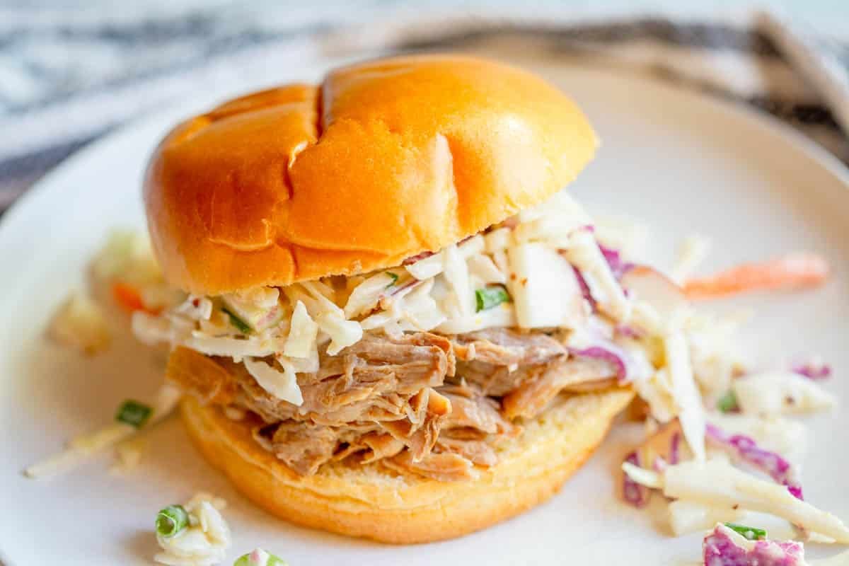 Slow Cooker Apple Cider Pulled Pork on a bun topped with apple cabbage slaw on a white plate.