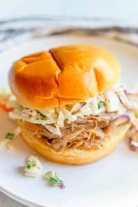 Slow Cooker Apple Cider Pulled Pork on a bun topped with apple cabbage slaw on a white plate.