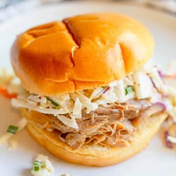 Slow Cooker Apple Cider Pulled Pork on a bun topped with apple cabbage slaw on a white plate.