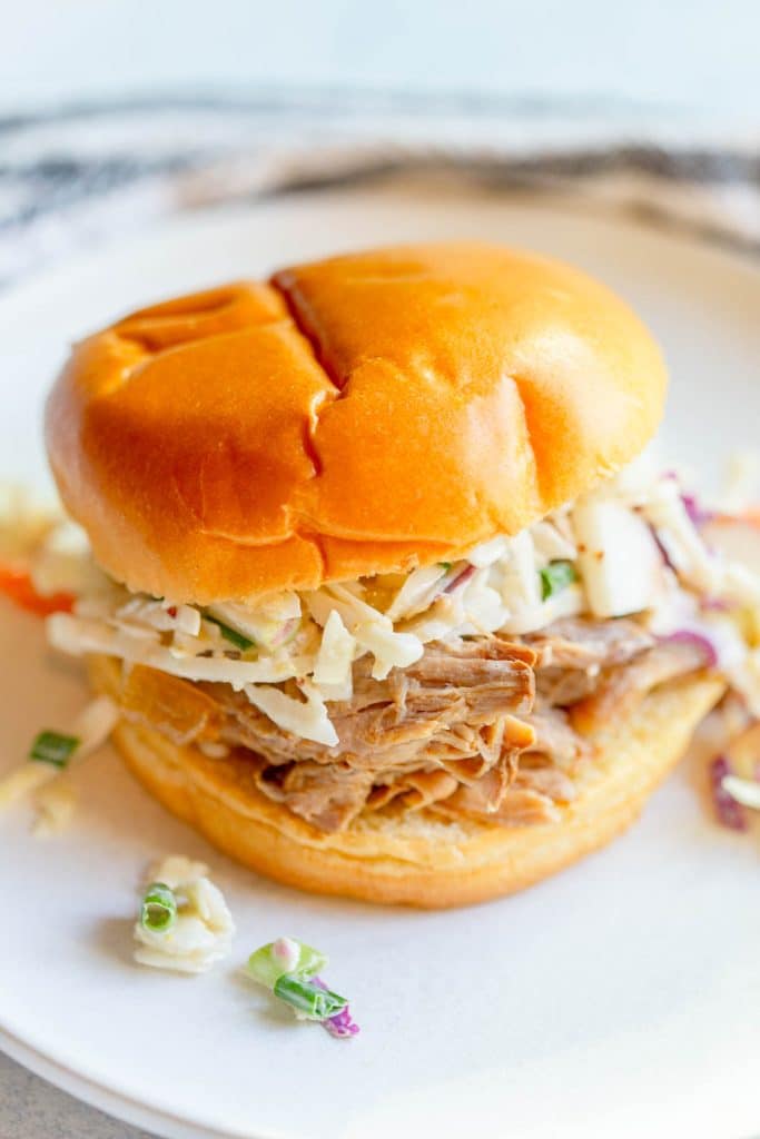 Slow Cooker Apple Cider Pulled Pork on a bun topped with apple cabbage slaw on a white plate.