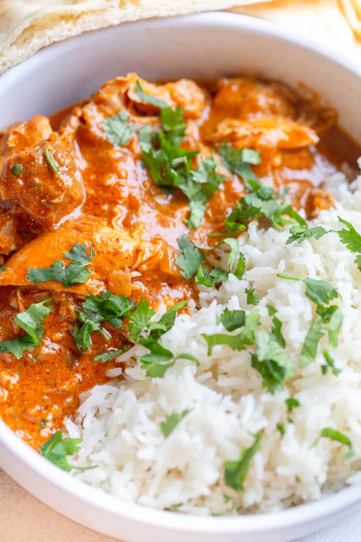 Slow Cooker Butter Chicken in a shallot cream colored bowl with white rice and chopped cilantro on top. There is a slice of naan in the background.