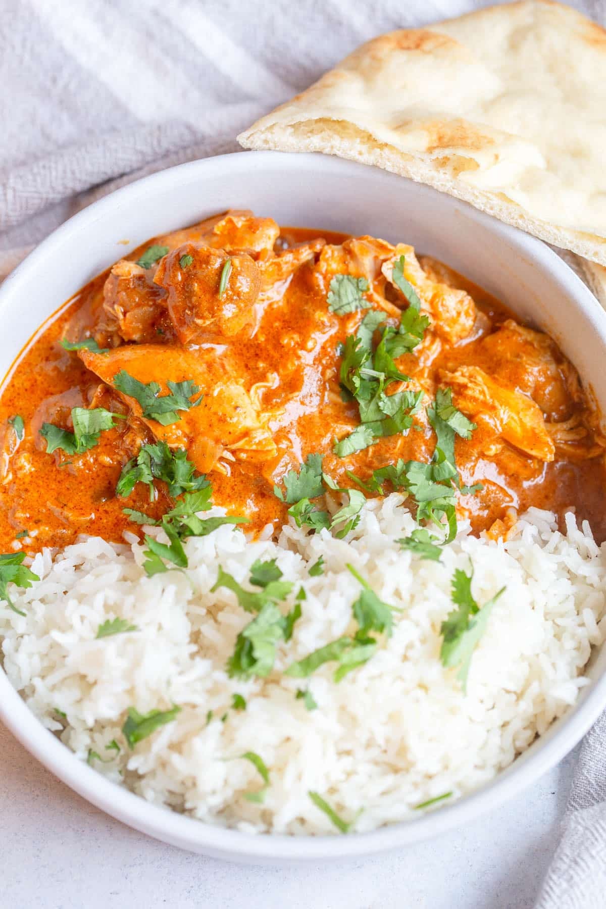 Slow Cooker Butter Chicken in a shallot cream colored bowl with white rice and chopped cilantro on top. There is a slice of naan in the background.
